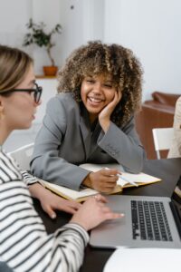 A Woman in Gray Blazer Smiling while Looking at Her Colleague Using a Laptop